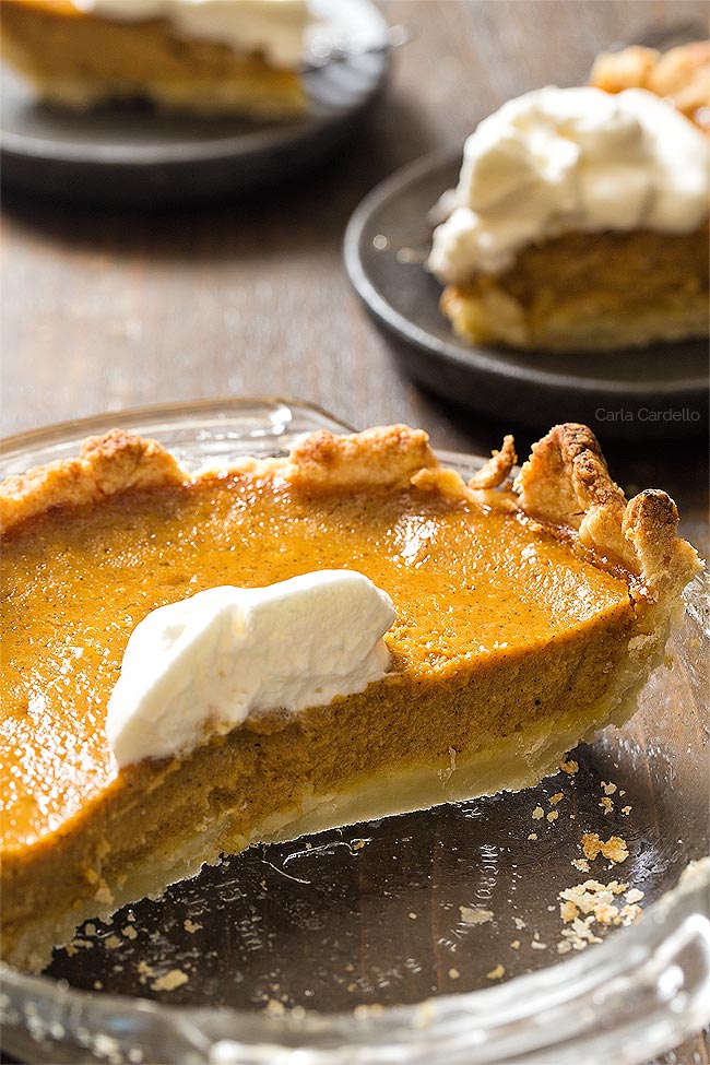 Half of pumpkin pie in glass pie pie with two slices on plates behind it