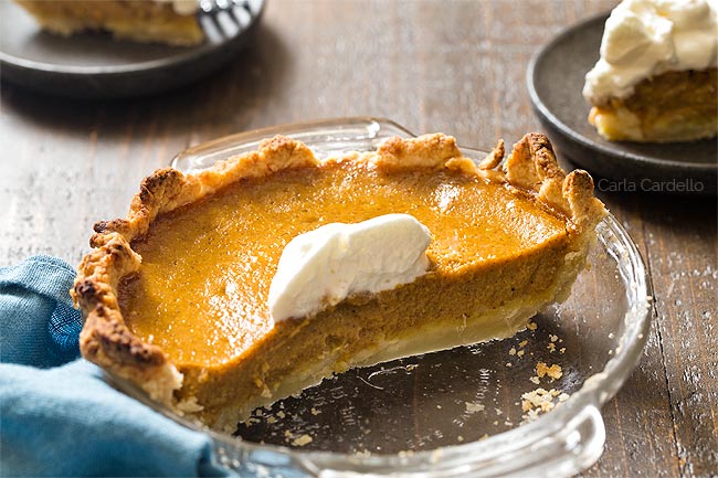 Half of pumpkin pie in glass pie pie with two slices on plates behind it