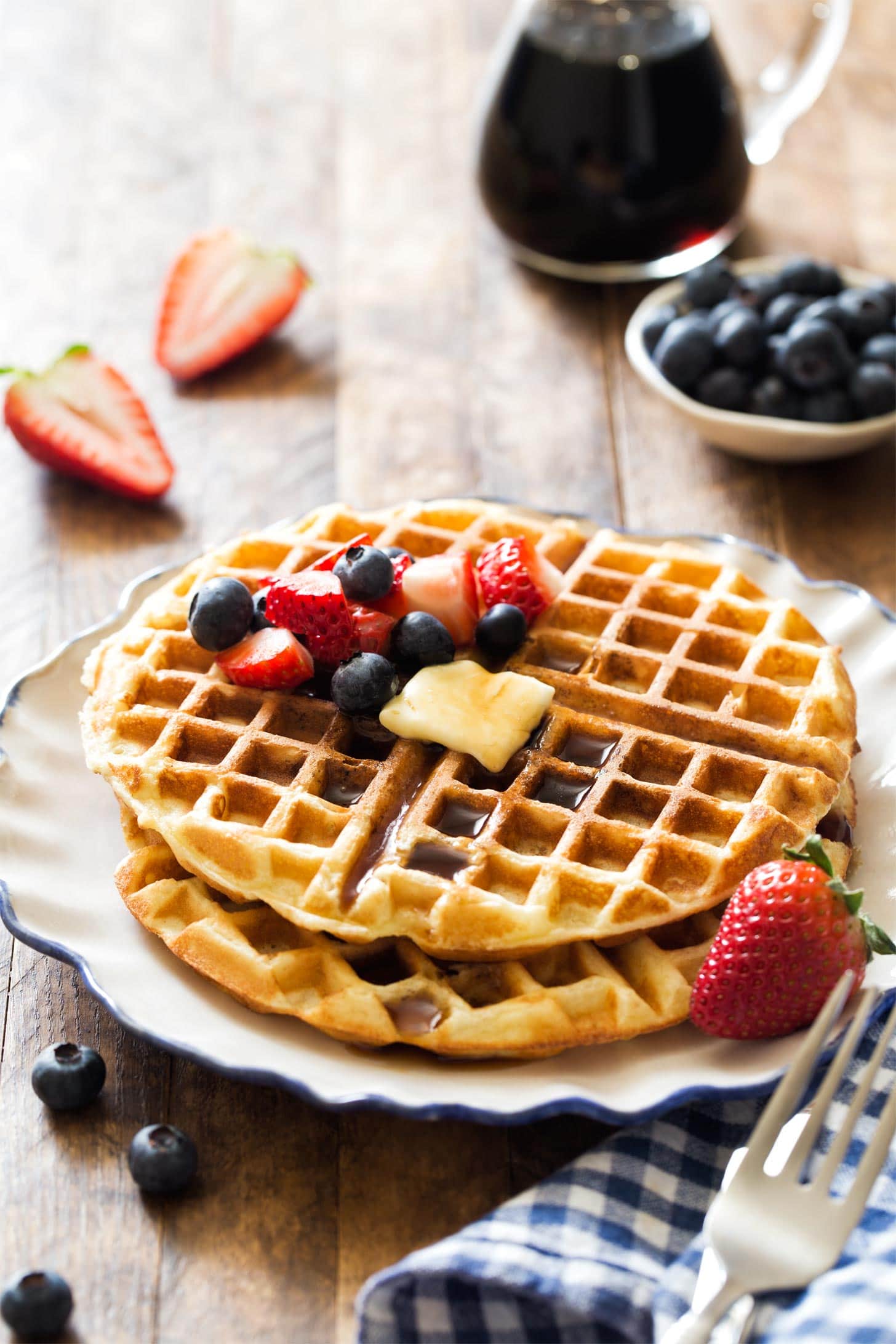 Stacked waffles for two with fresh berries, butter, and syrup on white plate